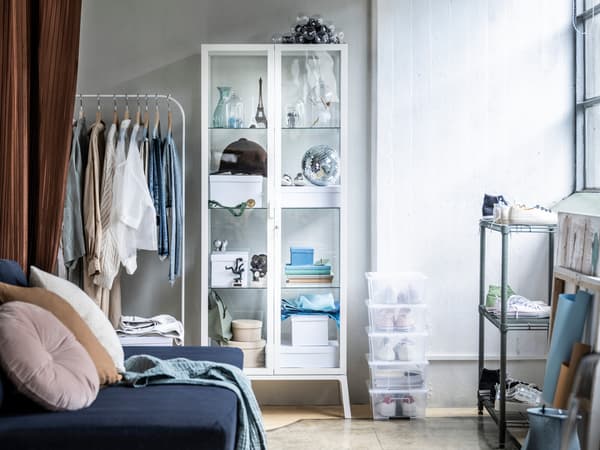 Bílý pokoj s modrou pohovkou a bílým šatním stojanem MULIG, u zdi stojí bílá vitrína MILSBO. Na podlaze leží několik průhledných krabic SAMLA.