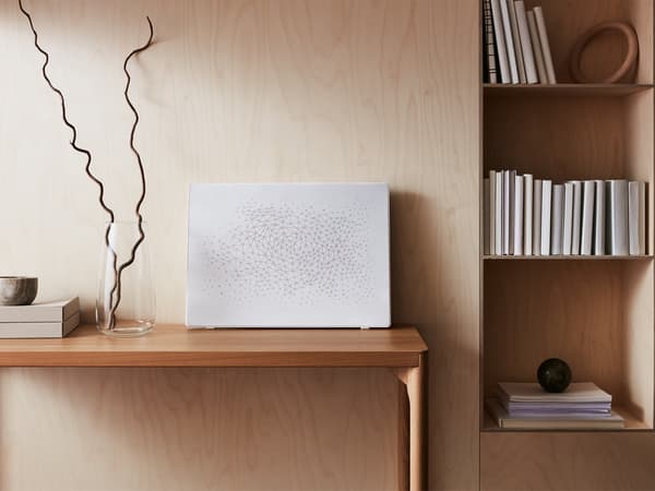 Bílý rám na obrazy SYMFONISK s reproduktorem WiFi, který stojí na stole s vázou vedle knihovny.