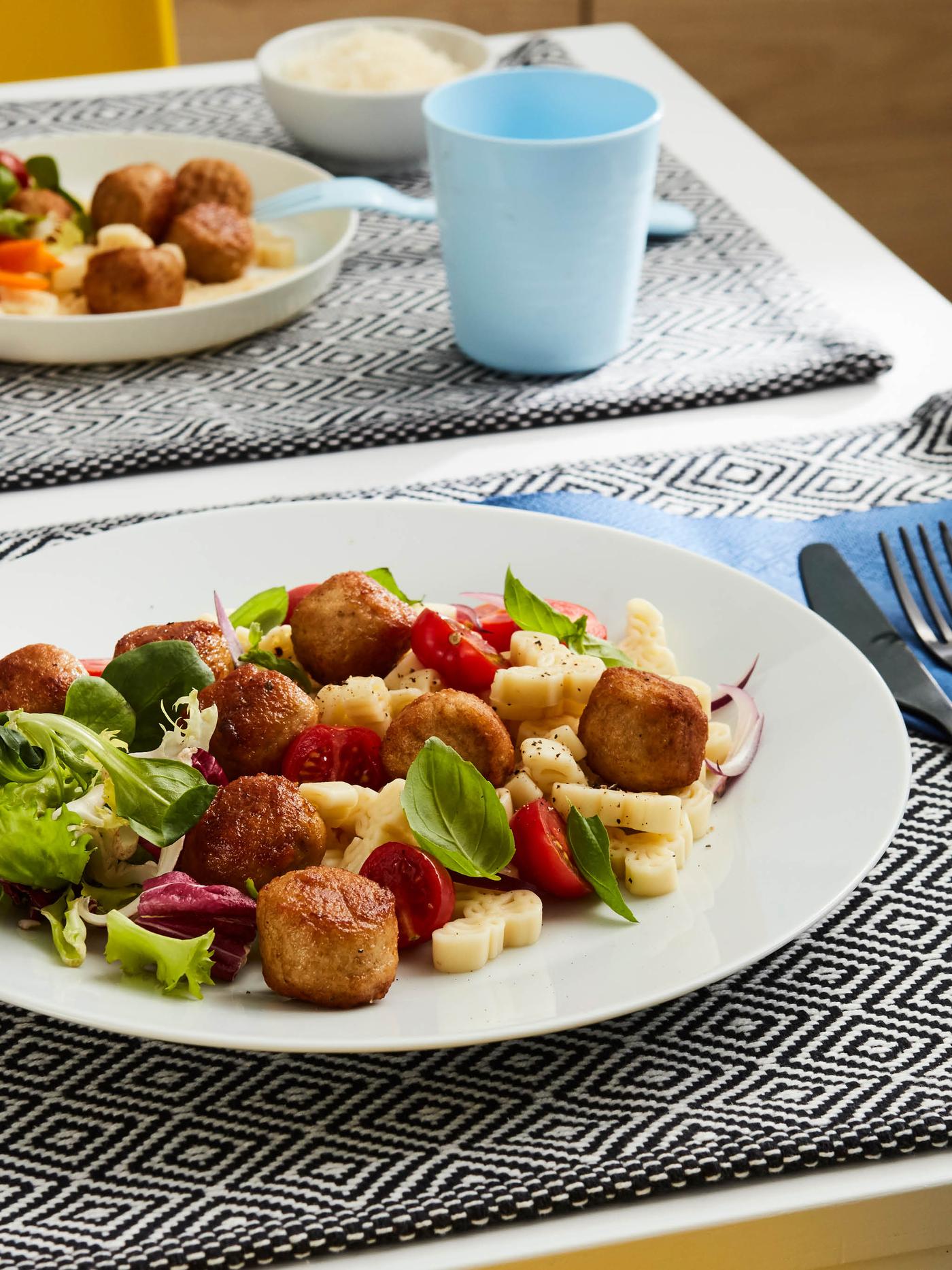 Bílý talíř s těstovinovým salátem s kuřecími kuličkami leží na černo-bílém prostírání, vedle talíře je na modrém ubrousku FANTASTISK černý příbor TILLAGD, v pozadí je na stejném prostírání krémový dětský talíř KALAS s malou porcí salátu, světle modrá vidlička a světle modrý hrnek KALAS.