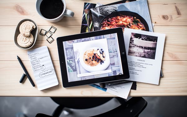 Bird’s-eye view of cooking inspiration on a tablet and in magazines, set at a table with coffee, note pad and pen.