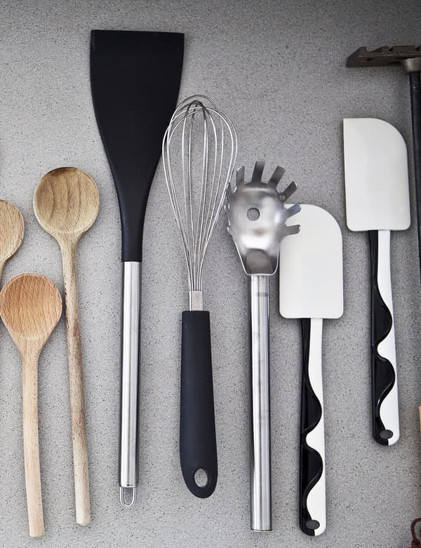 Bird's-eye view of kitchen utensils on a grey surface. Includes wooden spoons, and black and white spatulas.