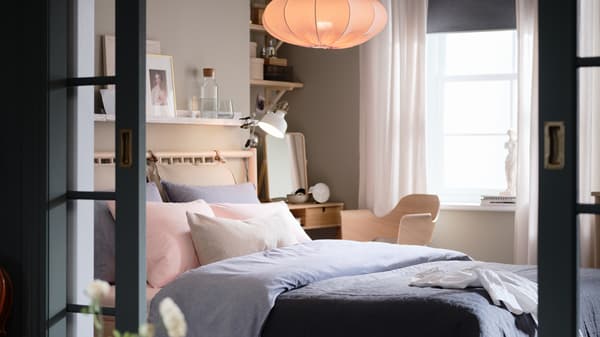 מיטת BJÖRKSNÄS מעץ ליבנה ושמיכה עם ציפת KOPPARBLAD בכחול כהה עומדת בחדר שינה מתחת למנורת תלייה.