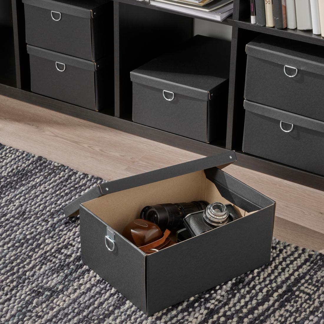 Black NIMM paper cardboard storage box on a floor carpet with its lid opened showing camera items being stored inside. A KALLAX shelf with multiple NIMM organisers stands in the background.  