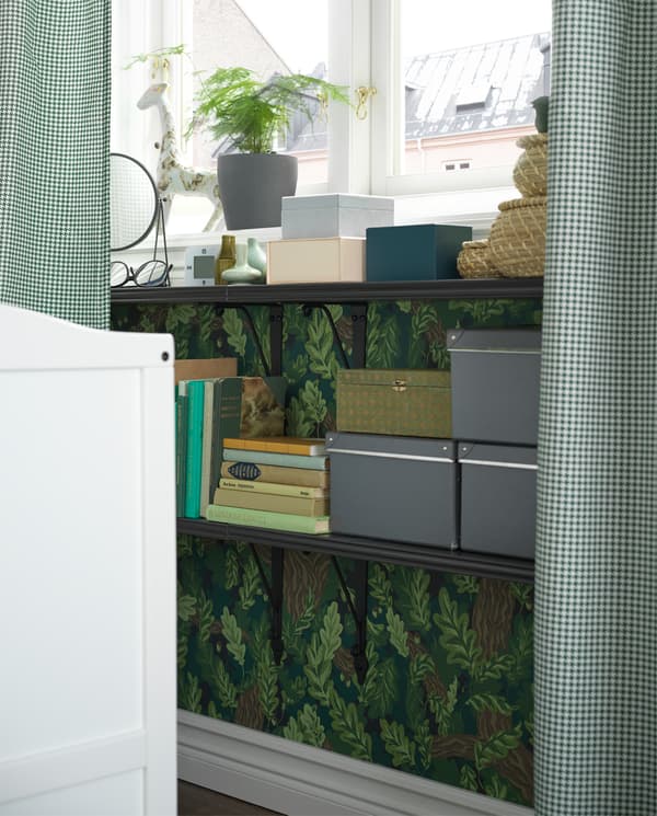 Black shelves are wall-mounted under a window and used as a bedside table. Books, boxes and decorative items stand here.