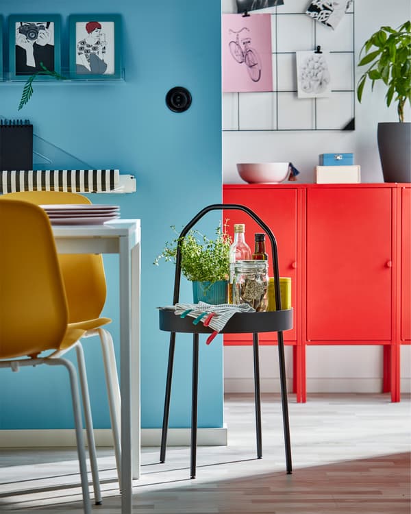 Black side table with handle, yellow chairs, white table, blue frames, red cabinet and a wall-mounted black memo board.