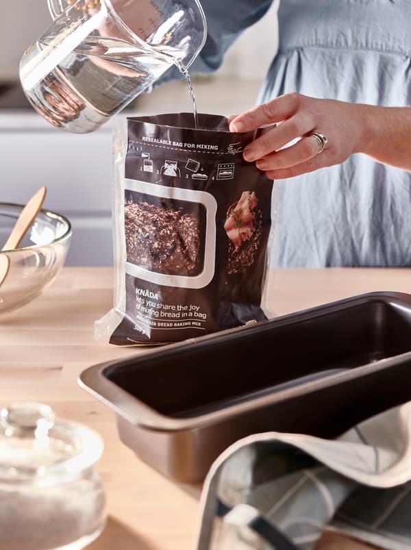 Blat de lucru de bucătărie cu o tavă de pâine HEMMABAK și alte accesorii de copt. Mâini care toarnă apă într-o pungă de amestec de copt KNÅDA.