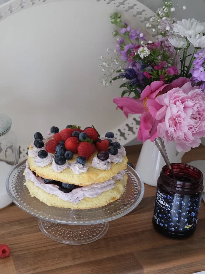 Blaubeer-Mascarpone Torte steht auf einem Kuchenständer auf der Küchenarbeitsfläche dahinter ein bunter Blumenstrauß und die Blaubeer Marmelade von IKEA.