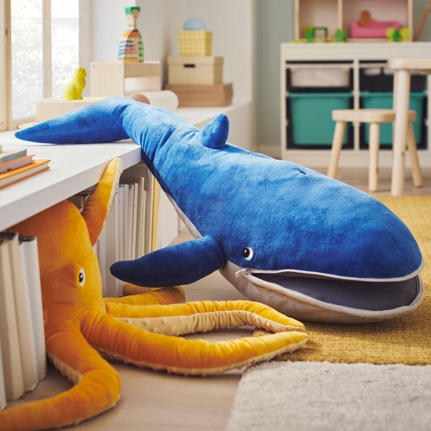 BLÅVINGAD-pehmolelumalliston sininen valas ja keltainen mustekala makaavat keltaisen matalanukkaisen LANGSTED-maton ja valkoisen kaapin päällä.