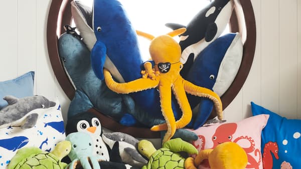 BLÅVINGAD soft toys are arranged in and in front of a round window with cushions in BLÅVINGAD ocean animal covers.