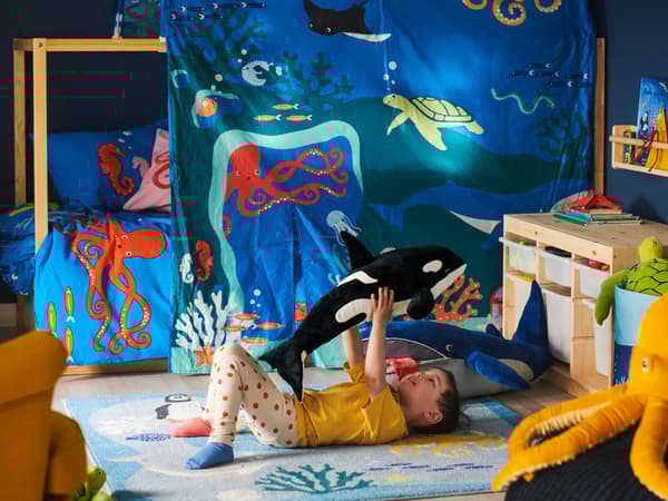 BLÅVINGAD szőnyegen fekvő kisfiú éppen felemel egy BLÅVINGAD orka puha játékfigurát. Mögötte egy BLÅVINGAD kék bálna fekszik a szőnyegen, a háttérben pedig egy kék BLÅVINGAD ágynemű és egy KURA baldachin látszik.
