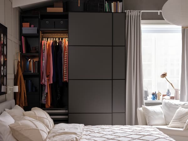 Blick auf ein Schlafzimmer mit einem dunkelgrauen PAX/MEHAMN Kleiderschrank. Eine seiner Türen ist geöffnet. Eine integrierte Beleuchtung erleuchtet die Kleidung auf der Kleiderstange des Schrankes.