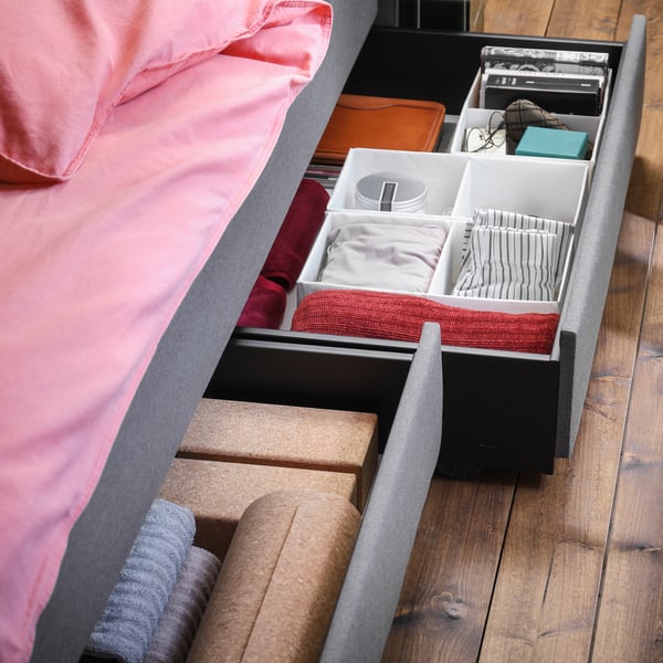 Blick auf eine Seite eines grauen HAUGA Bettgestells mit zwei breiten Bettkästen, die ausgezogen sind und in denen sich Textilien und Fächer mit Accessoires befinden.