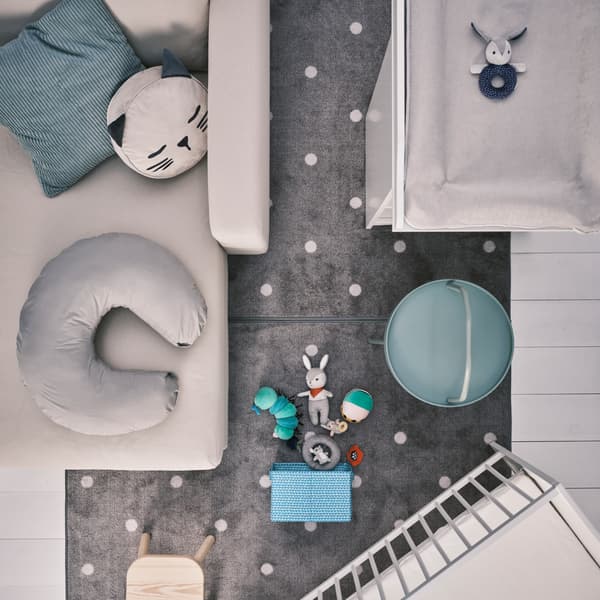Blick auf einen gepunkteten/grauen LEN Teppich auf weißem Boden, u. a. mit SMÅGÖRA Wickeltisch, Babybett und Babyspielzeug.