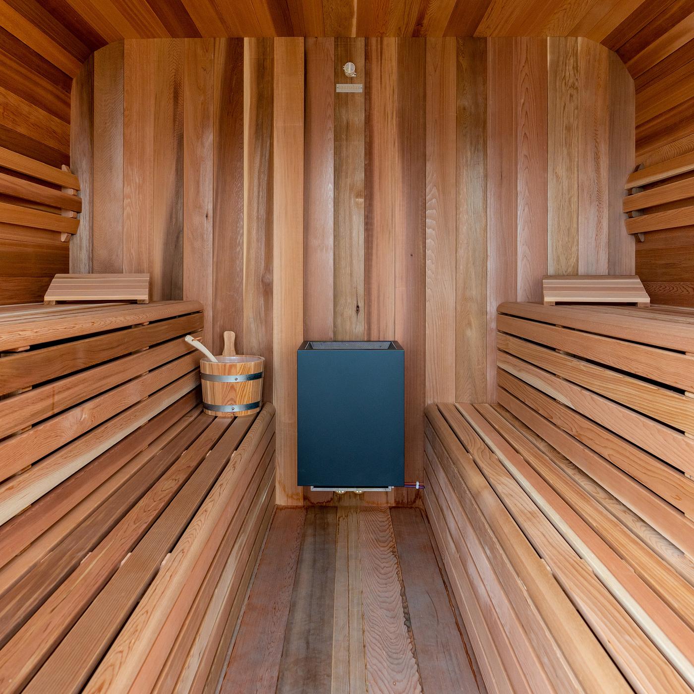 Blick in einen Saunacube, der mit hellem Holz und mehrstufigen Sitzbänken auf beiden Seiten eingerichtet ist. An der hinteren Wand steht der Sauna-Ofen