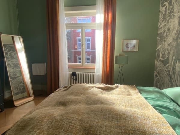 Blick über ein gemachtes Bett zum Fenster des Schlafzimmers mit langen Vorhängen. Daneben steht ein grosser Standspiegel. Auf der anderen Seite eine kleine Stehlampe mit rundem Lampenschirm. 