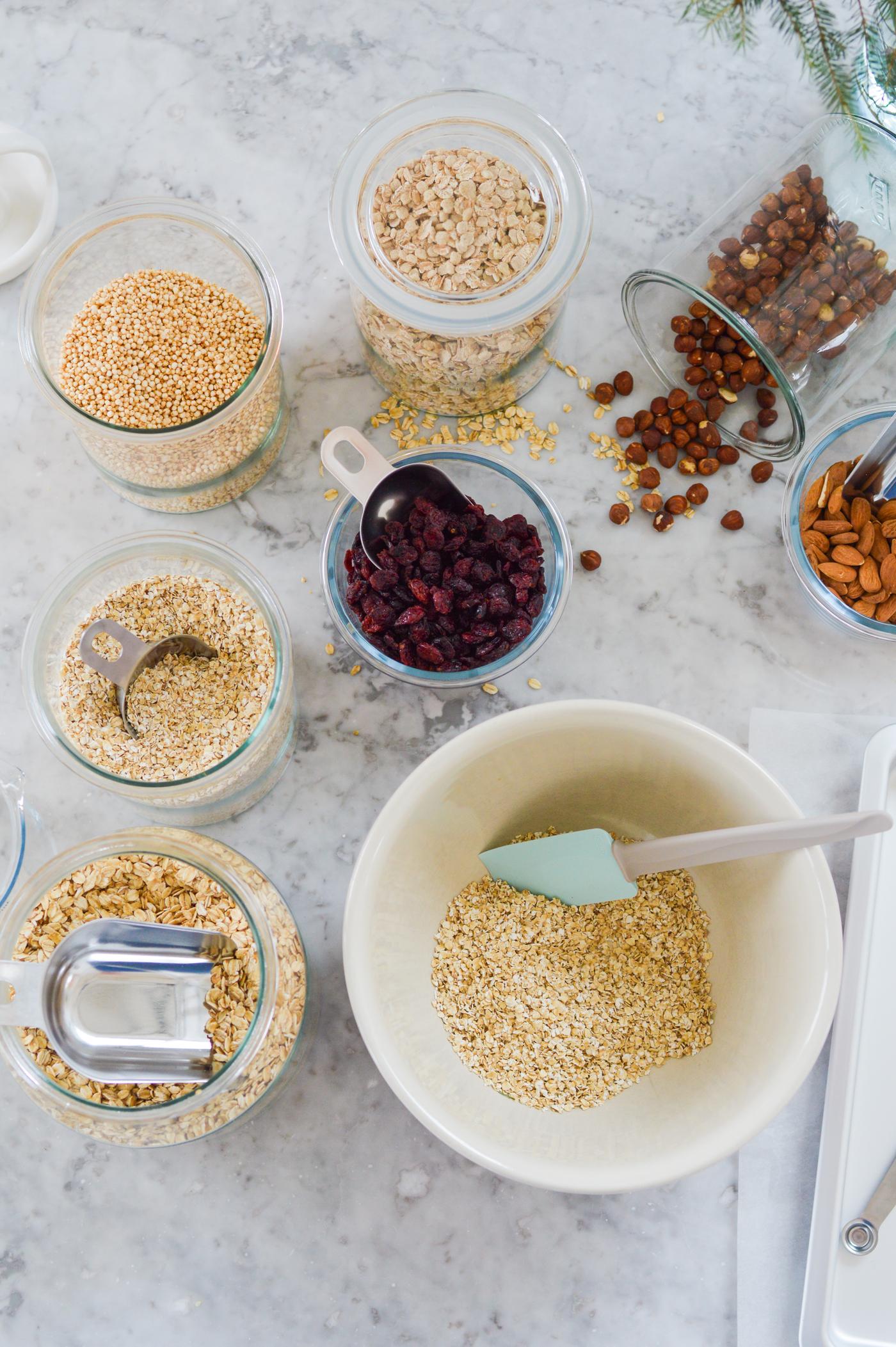 Blick von oben auf verschiedene Getreidesorten, Nüsse und getrocknete Beeren in Schalen und Gläsern auf einer Marmoroberfläche. In der Mitte steht eine Rührschüssel mit Getreide und einem Spatel.