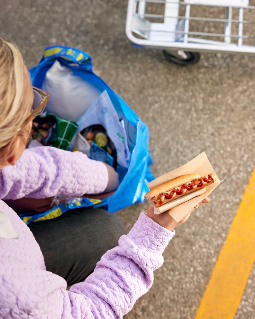 Blonde woman digging through an IKEA FRAKTA bag while holding an IKEA hotdog with ketchup.