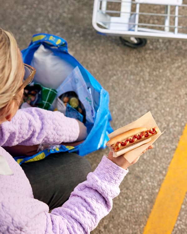 Blonde woman digging through an IKEA FRAKTA bag while holding an IKEA hotdog with ketchup.