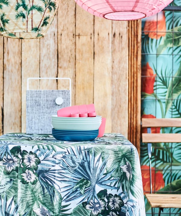 Blue and green leaf fabric covering a table, a stack of blue plates, a white speaker and a patio chair plus hanging lanterns.