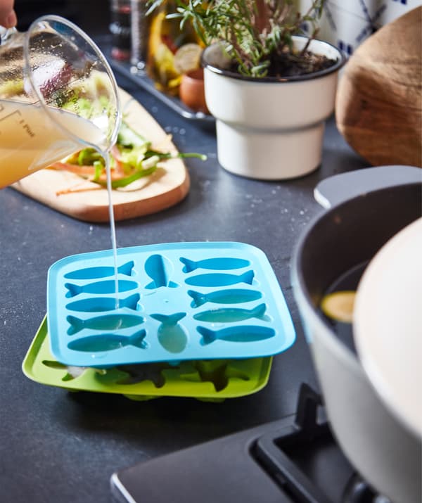 Blue and green PLASTIS ice-cube trays on a kitchen worktop. The top one is being filled with broth poured from a glass beaker.