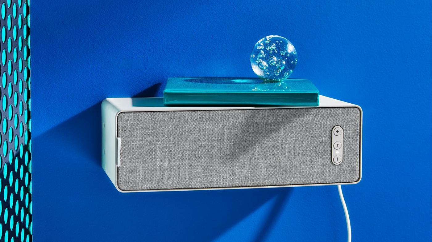 Bluetooth reproduktor pripevnený na modrej stene, na ktorom je položená modrá kniha a sklenená dekoračná guľa.