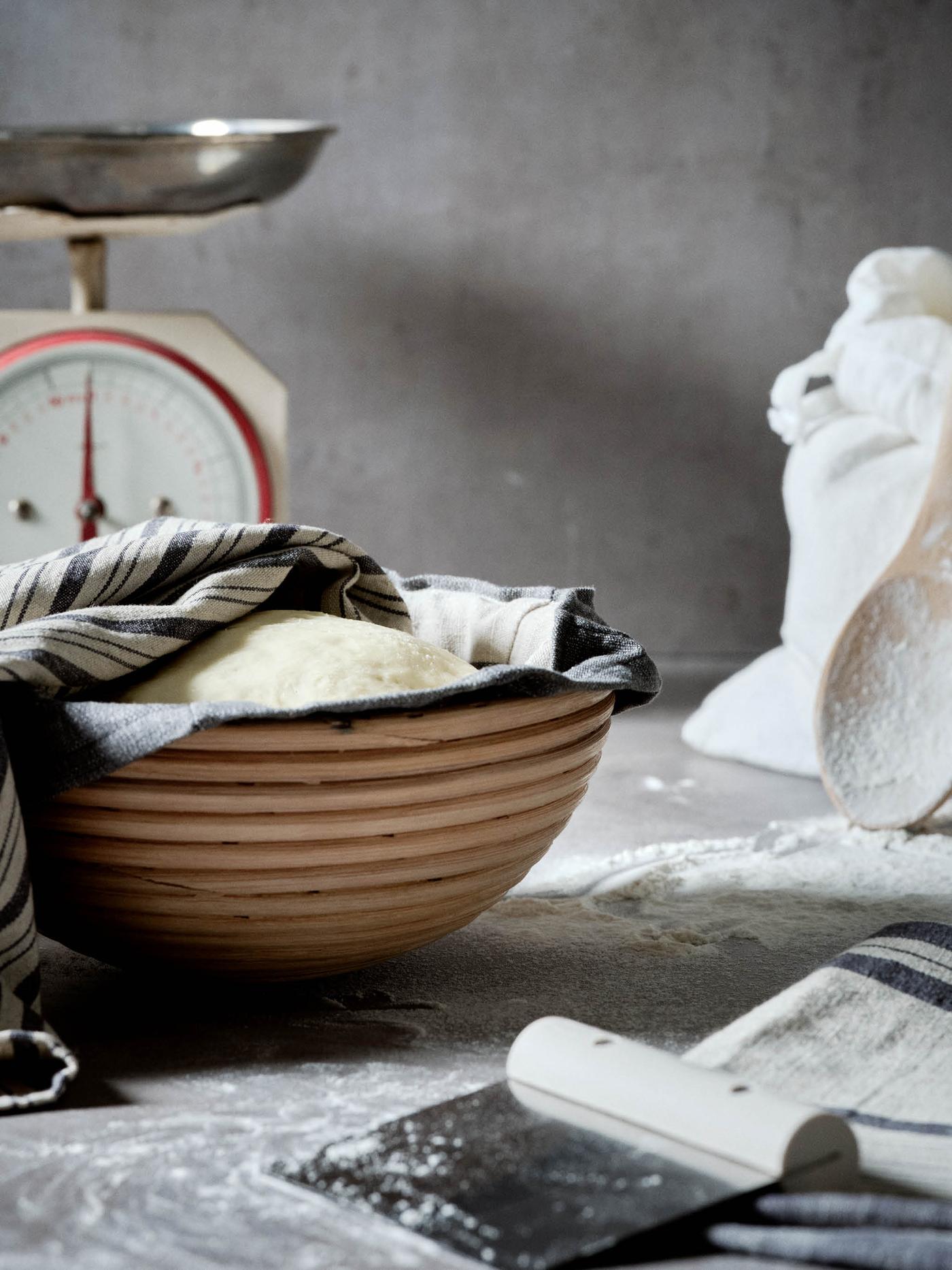 Bochánek těsta kyne v košíku na kynutí JÄSNING, v pozadí je váha, pytlík mouky a lžíce RÖRT, v popředí leží utěrka MARIATHERES a kráječ na těsto LÄTTBAKAD.