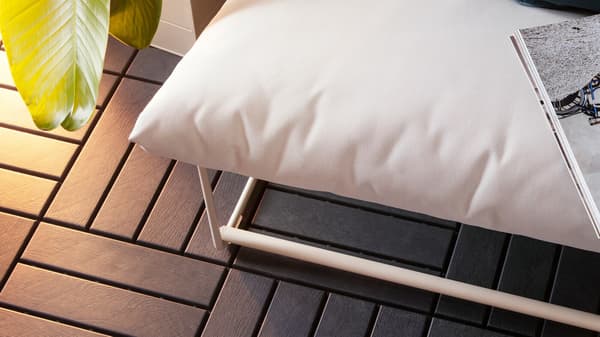 Bodenrost in Holzoptik unter einem Gartensofa mit weißen Polstern. Darauf ist ein Buch zu sehen.