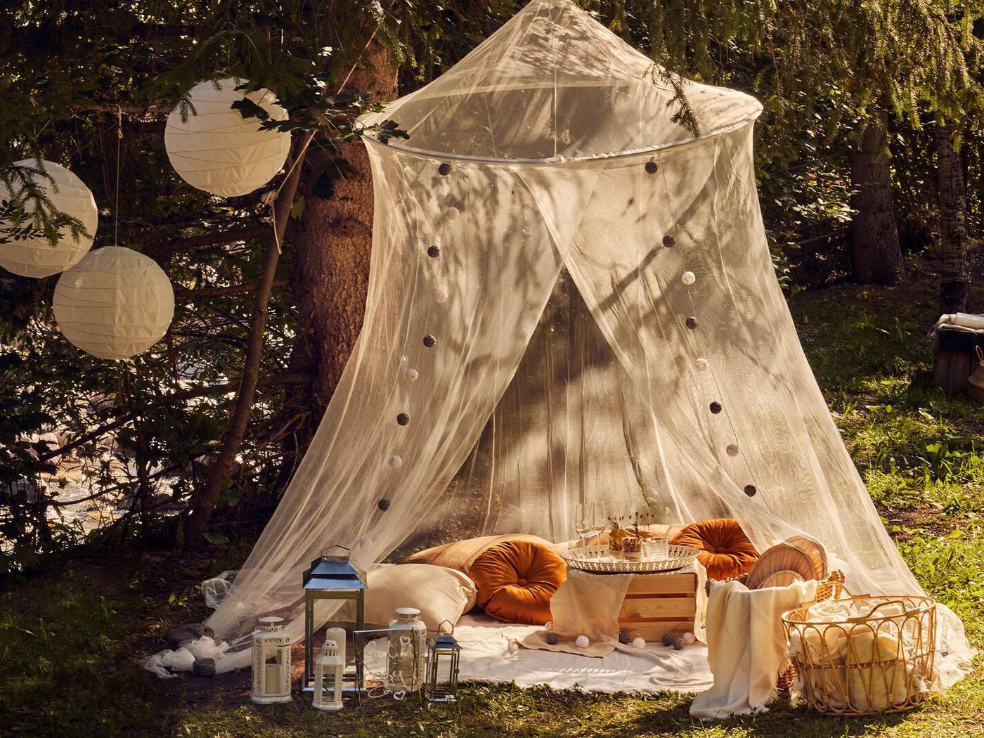 Boho Hochzeitszenerie. Es ist eine Picknick-Sitzecke mit einem weißen Baldachin zu sehen. Lichterketten, Reispapierleuchten, einer Decke mit vielen Kissen und Laternen befinden sich drumherum. 