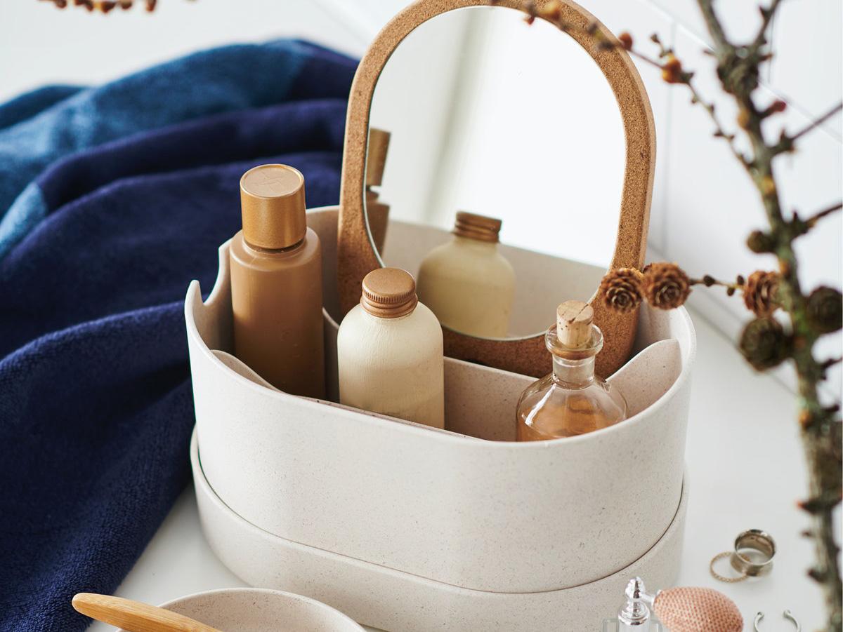 Boite de rangement ovale avec trois bouteilles de produits cosmétiques et un miroir avec un contour façon liège posé sur une table blanche avec une serviette bleu en fond et à côté des bagues , un parfum et une petite plante