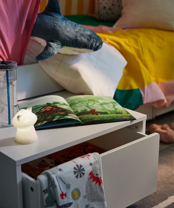 Boîte de rangement tirée de dessous le lit et tenant lieu de table de chevet, sur laquelle sont posés une veilleuse, un livre et une bouteille d'eau.
