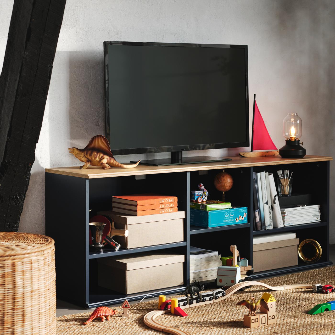 Boîtes, livres et jouets sur les tablettes ouvertes d’un banc TV SKRUVBY bleu noir, sur lequel se trouve un téléviseur noir.