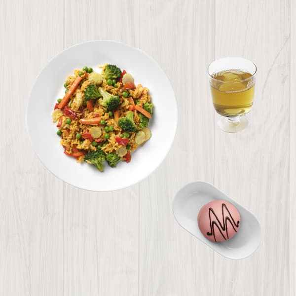 Bol alb cu orez prăjit cu legume (broccoli, morcovi, mazăre, ardei roșu), alături de un pahar cu suc galben și un desert roz decorat cu ciocolată pe farfurie albă.