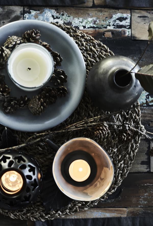 Bol gris avec bougie blanche dans un verre et pommes de pin posées sur un set de table tissé.