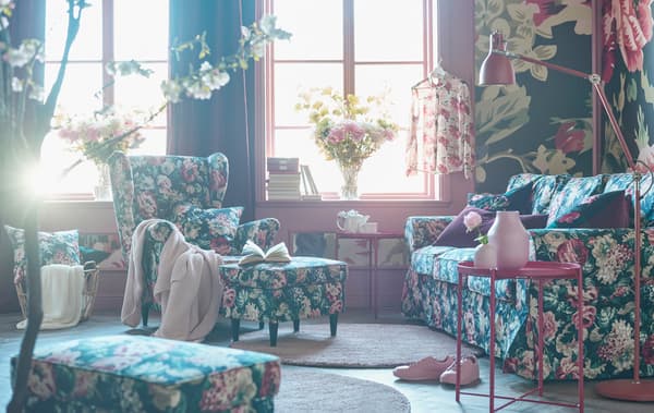 Bold floral patterned sofa, armchair, footstool and cushions in a living room with a bold floral patterned wallpaper.