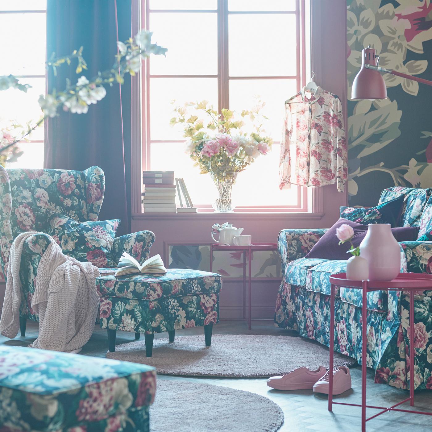 Bold floral patterned sofa, armchair, footstool and cushions in a living room with a bold floral patterned wallpaper.