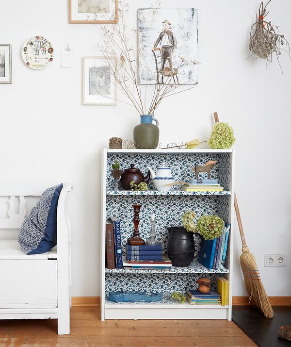 Books and ornaments on a small bookcase with floral fabric interior, in a room with white walls and wooden floor.