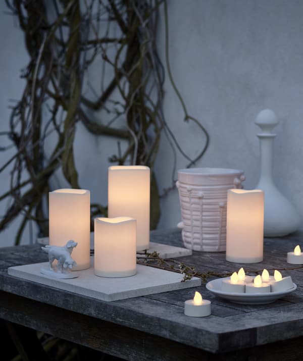Bougies à DEL de tailles variées déposées sur une table à l’extérieur, près d’un mur blanc.