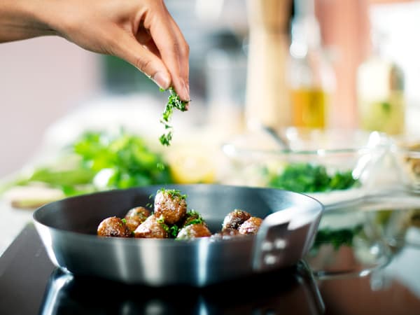 Boulettes végétales HUVUDROLL en train d’être frites dans une poêle noire sur une table de cuisson à induction. Une main les parsème de persil haché.