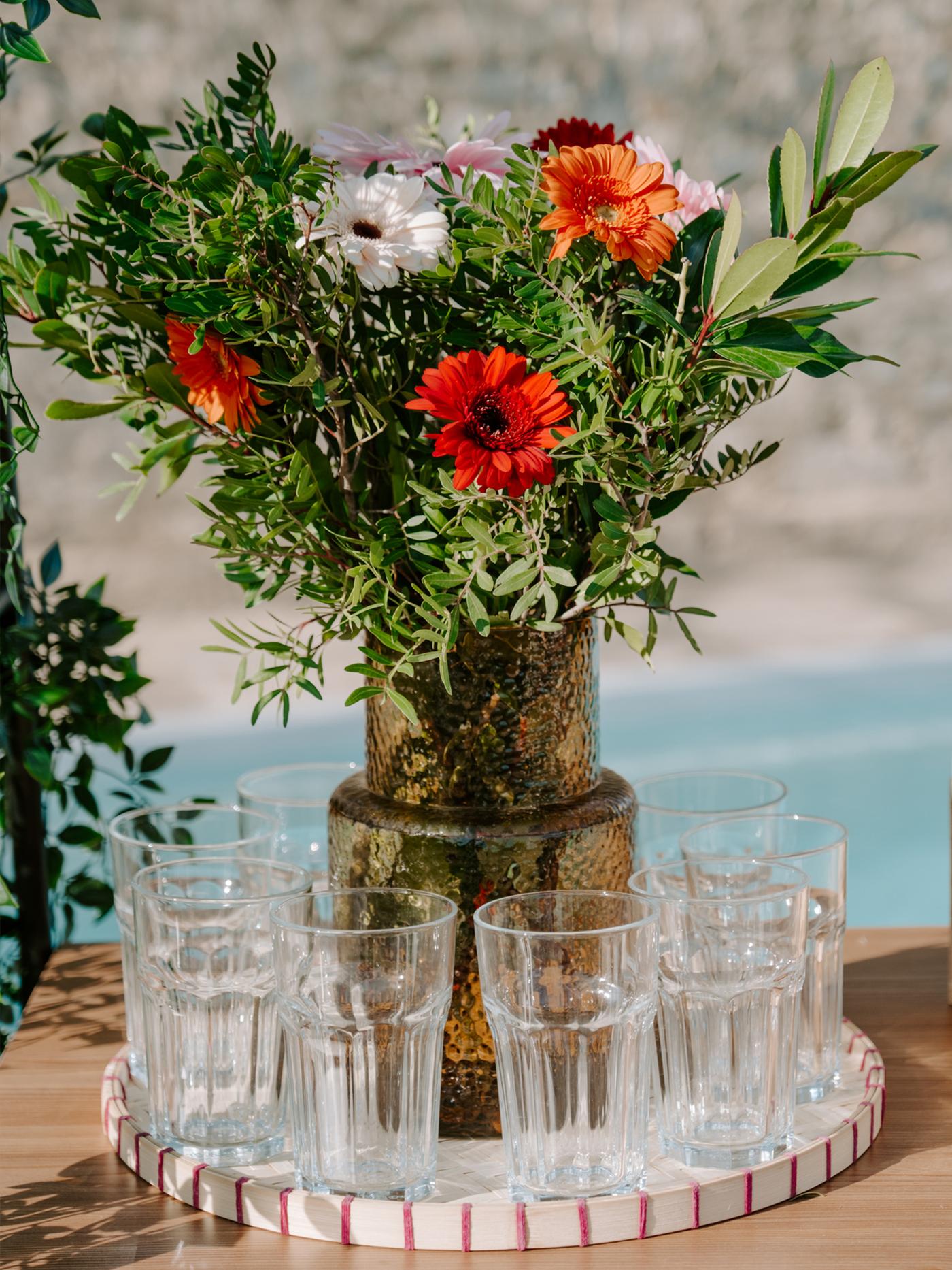 Bouquet de fleurs dans un vase en verre coloré avec des grands verres diposés autour sur un plateau rond blanc et rouge