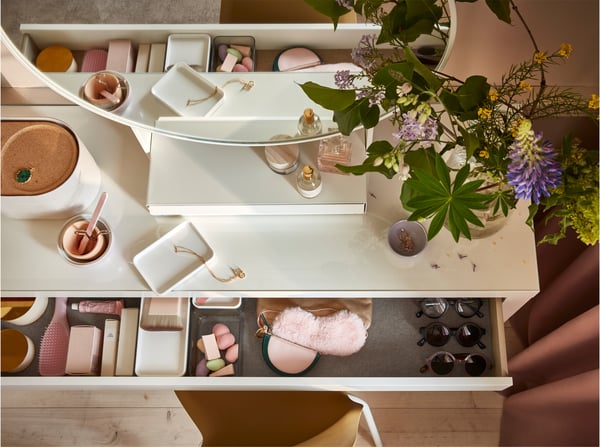 Bovenaanzicht van een witte toilettafel en een open lade waarin make‑upsponsjes, zonnebrillen en roze accessoires liggen.