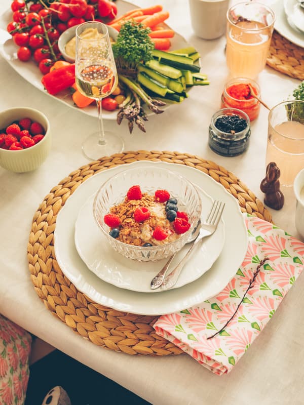 Bowl of granola with berries on white plates, placed on a woven placemat with a floral napkin on a table set with fruits and drinks.