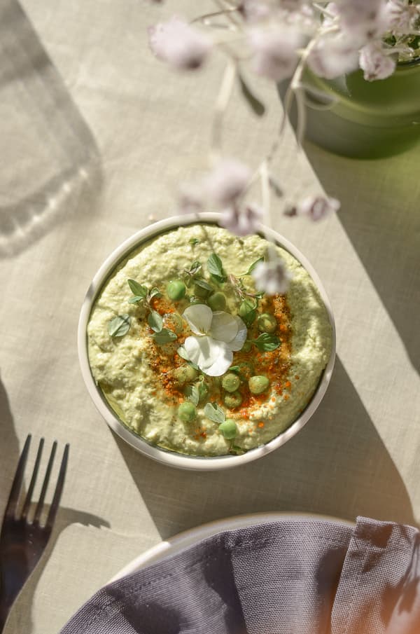 Bowl of green dip with peas, microgreens and flower on light tablecloth, fork and napkin in foreground.
