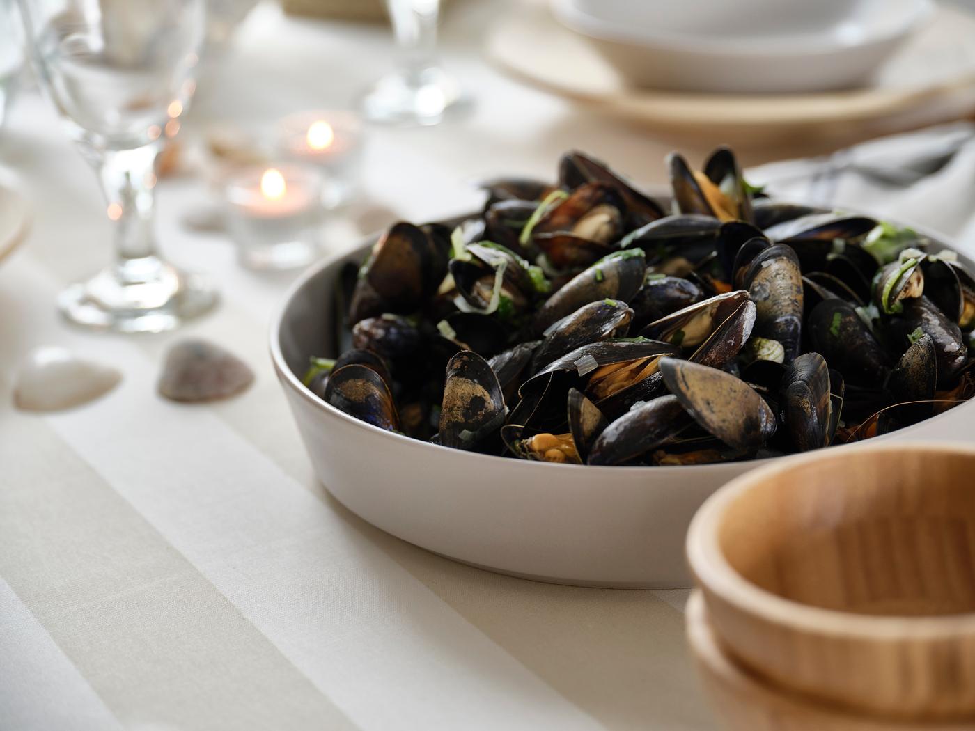 Bowl of mussels on a softly set table with glasses, tealights and a wooden serving bowl nearby.