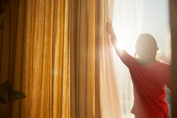 Boy wearing a red t-shirt opening up yellow curtains to let the sun in.