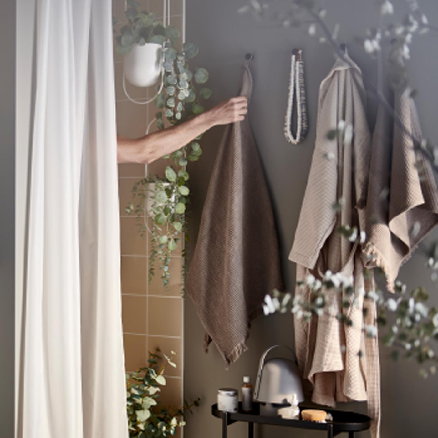 Braço de homem a alcançar uma toalha em castanho de trás de cortina de duche BASTSJÖN, em branco, numa casa de banho tipo spa acolhedora com algumas plantas.