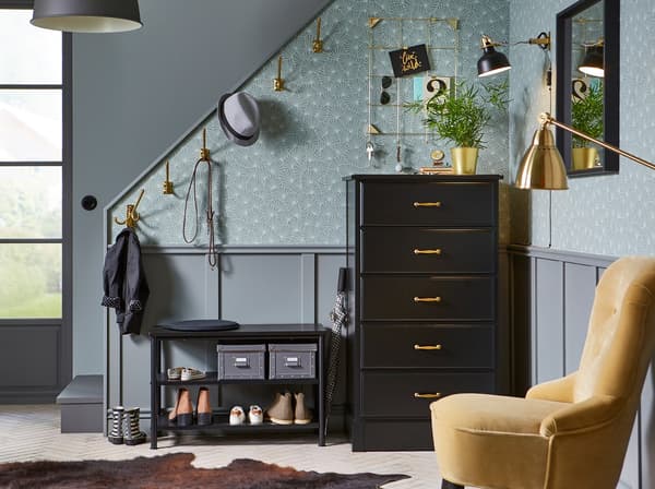 Brass coloured KÄMPIG hooks, a black PINNIG bench and UNDREDAL chest of drawers arranged against a grey wallpaper.