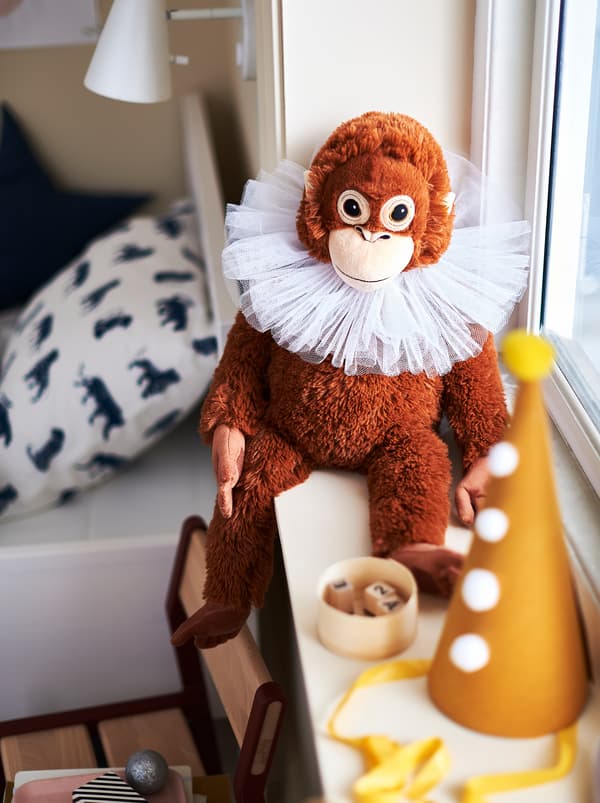 Brązowa pluszowa małpka DJUNGELSKOG i jej złota czapka leżące na parapecie okna w pokoju dziecięcym urządzonym tak, aby przypominał namiot cyrkowy.