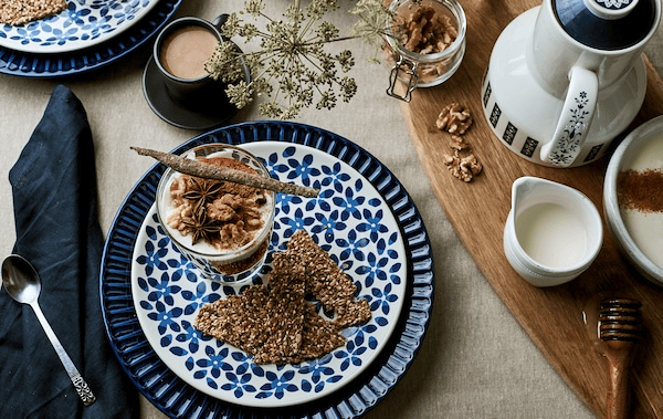 Breakfast table set with linen, patterned blue plates, granola in a glass and coffee accessories on a chopping board.