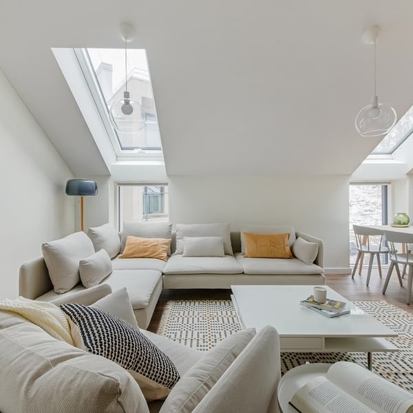 Bright and airy attic living room with a modern Scandinavian design. A large L-shaped beige sectional sofa is decorated with neutral and mustard yellow cushions. A white coffee table with a magazine and mug sits on a geometric-patterned rug. Skylights and tall windows fill the room with natural light. To the side, a cozy armchair with a black-and-white pillow and a cream throw faces the sofa. In the background, a small dining area with pale gray chairs and a light wood table is visible. The space is clean, minimalist, and inviting.