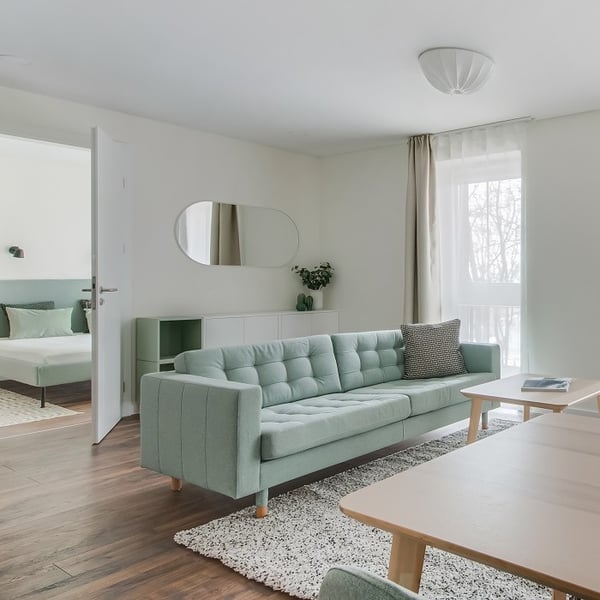 Bright and airy modern living room with a pastel green upholstered sofa centered on a textured white rug. A rectangular wooden coffee table sits in front of the sofa. On the left, a doorway opens to a bedroom with a matching green bed frame. The room features a white wall-mounted cabinet, a round-edged mirror, and a small green vase with greenery. Sheer white curtains with beige drapes allow natural light to filter through a large window, enhancing the soft and minimalist Scandinavian-style décor.